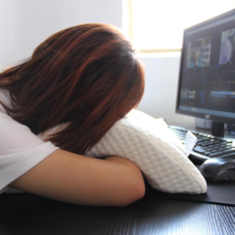 Person resting head on white memory foam pillow at desk with computer, ergonomic support for neck and arms